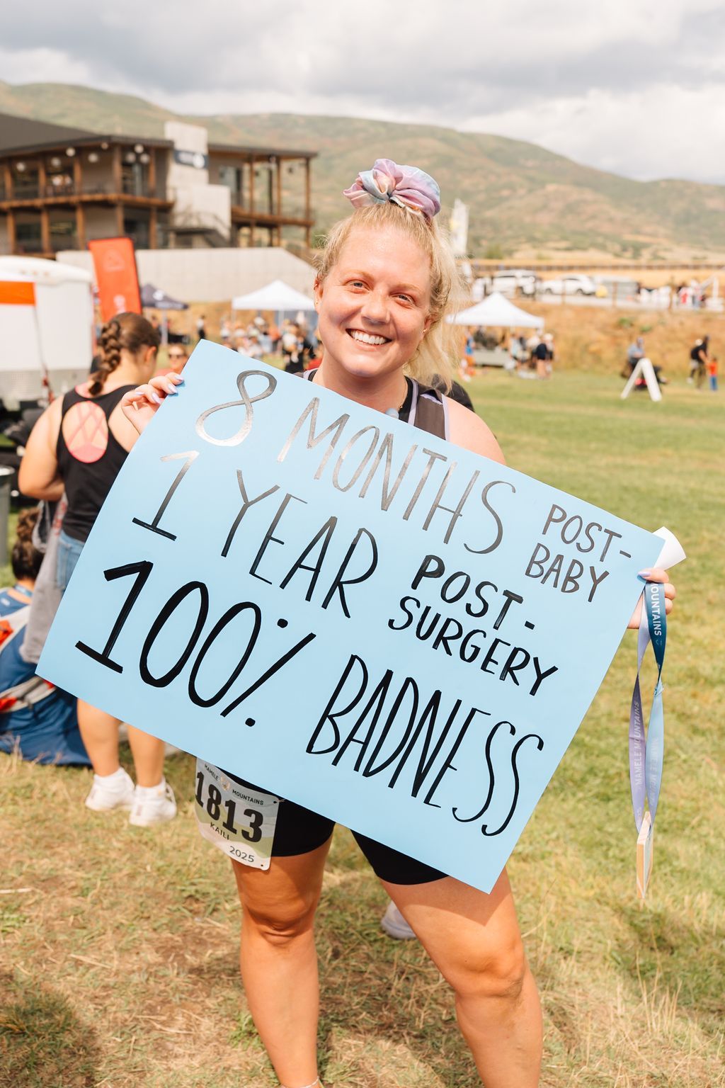 A racer holding a handmade sign reading nine months post baby, one year post surgery, one hundred percent badass.